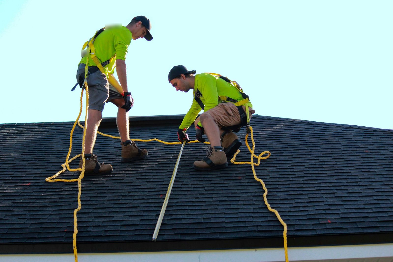 Two installers in safety harnesses measuring a shingle roof